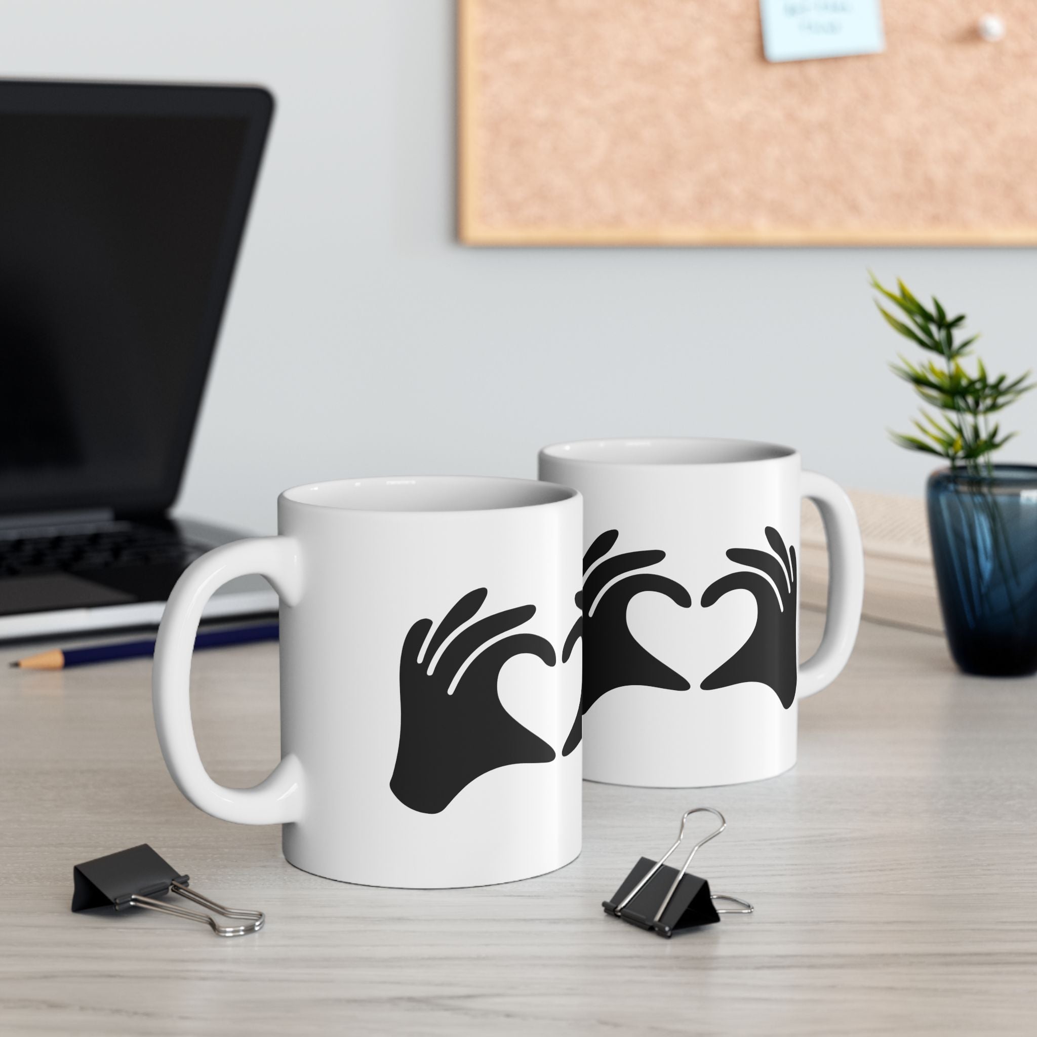 Two white mugs with black hand designs on a desk with a laptop and plant in the background.
