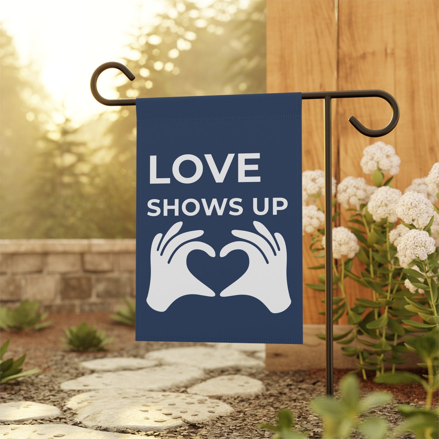 Garden flag with 'LOVE SHOWS UP' text and hands holding a heart, displayed outdoors.