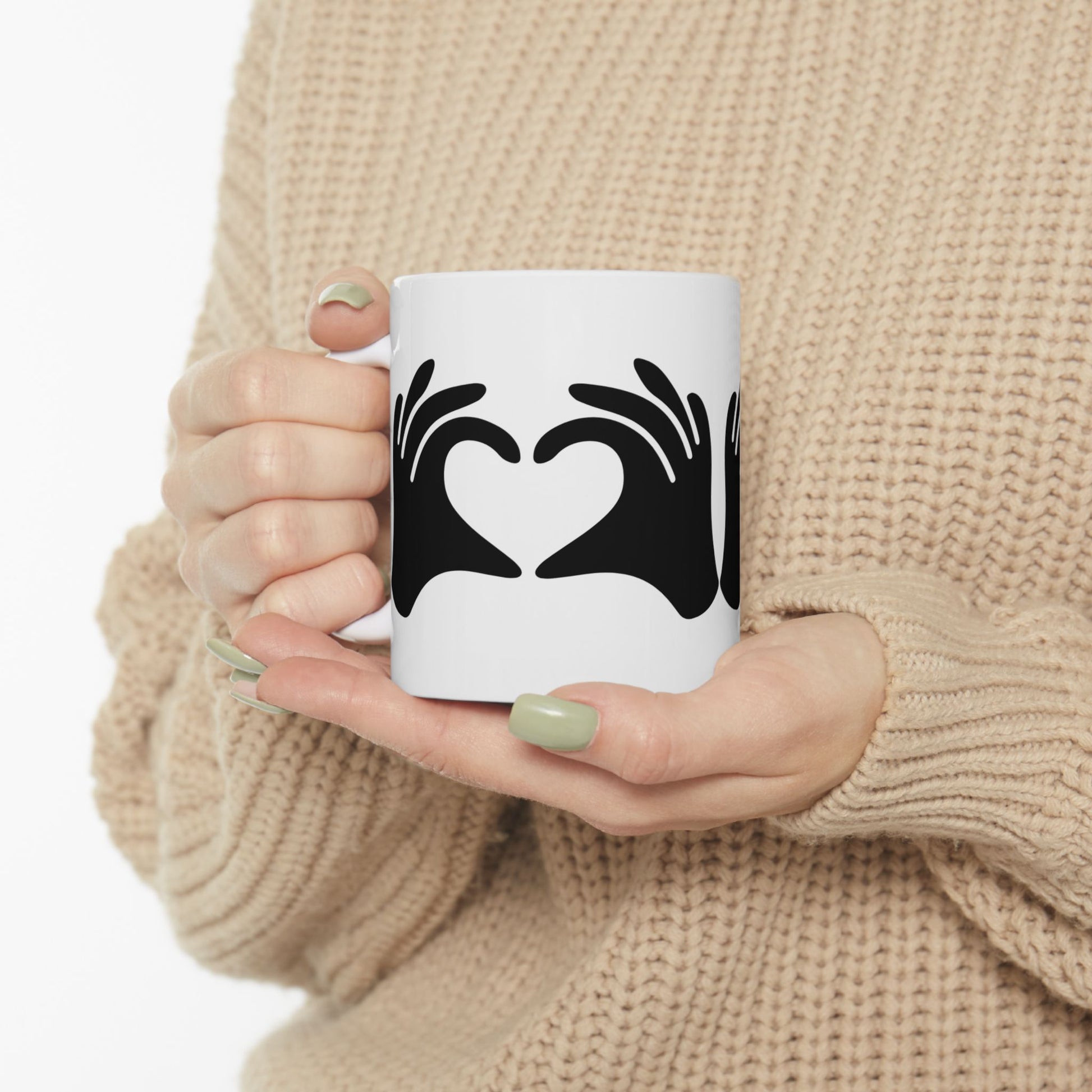 Person holding a white mug with black design of hands forming a heart, wearing a beige sweater.